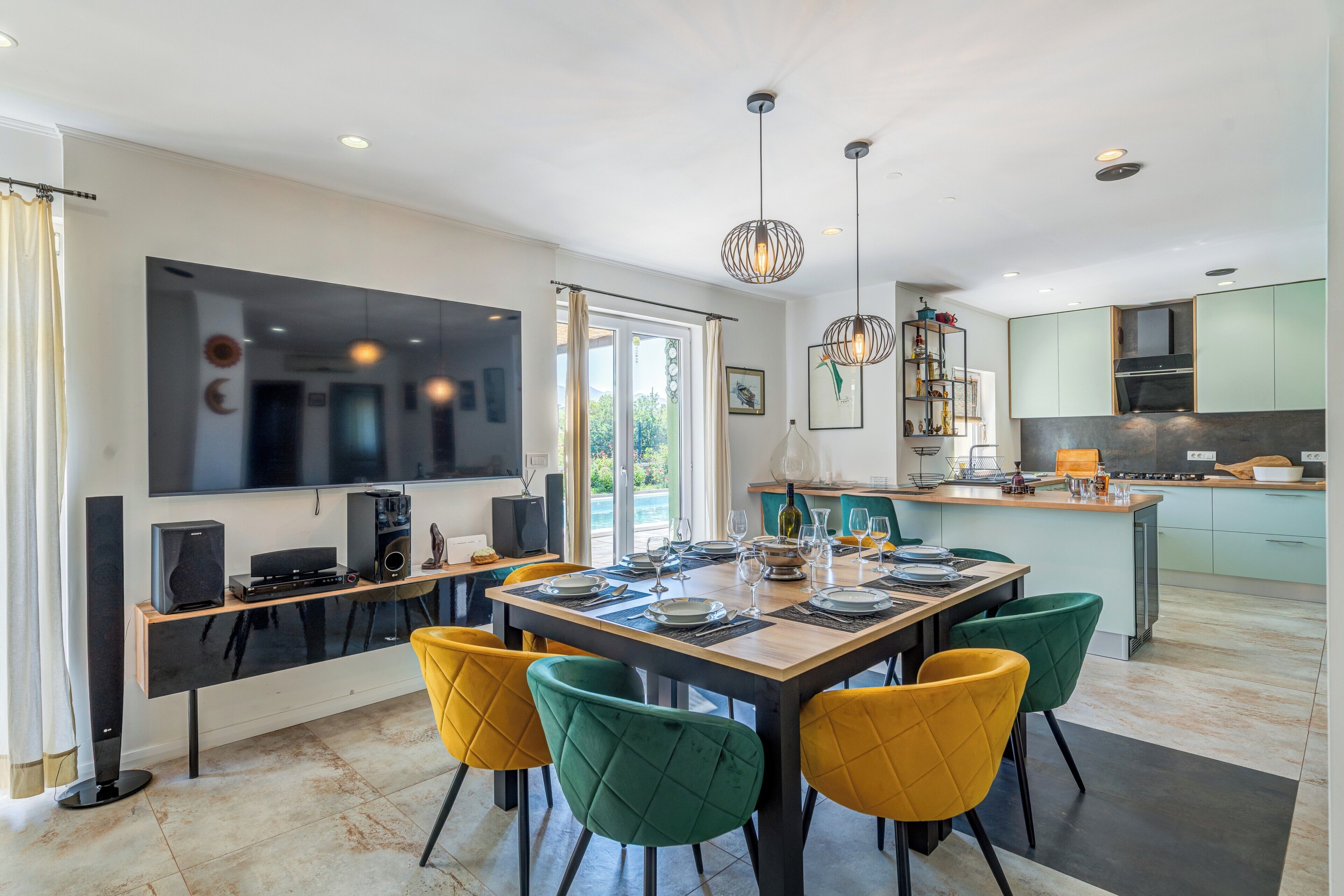 A modern kitchen with a dining area, featuring a large TV, a dining table set for six, and a kitchen counter with appliances.