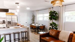 A modern kitchen with a stainless steel refrigerator, a dining area with a table and chairs, a brown leather sofa, and a large green plant.