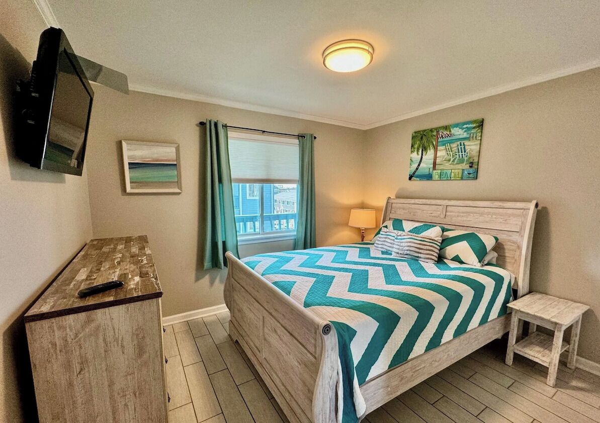 Une chambre à coucher avec un lit, une table de chevet, un téléviseur fixé au mur, une fenêtre avec des rideaux et un tableau accroché au mur.