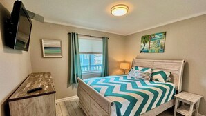 Une chambre à coucher avec un lit, une table de chevet, un téléviseur fixé au mur, une fenêtre avec des rideaux et un tableau accroché au mur.