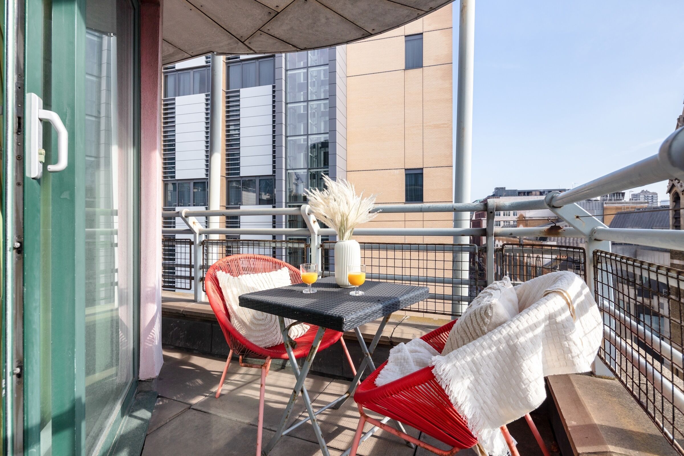 Un balcone con poltroncine in vimini rosso, un tavolino con un vaso e una bevanda arancione, e vista su edifici moderni.