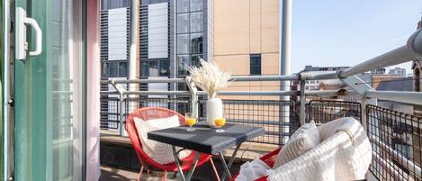 Un balcone con poltroncine in vimini rosso, un tavolino con un vaso e una bevanda arancione, e vista su edifici moderni.