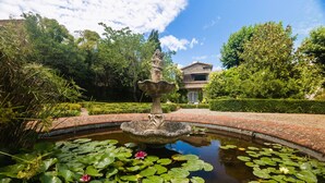 Ein Garten mit einem Brunnen, einem Teich mit Seerosen und einem Gebäude im Hintergrund.
