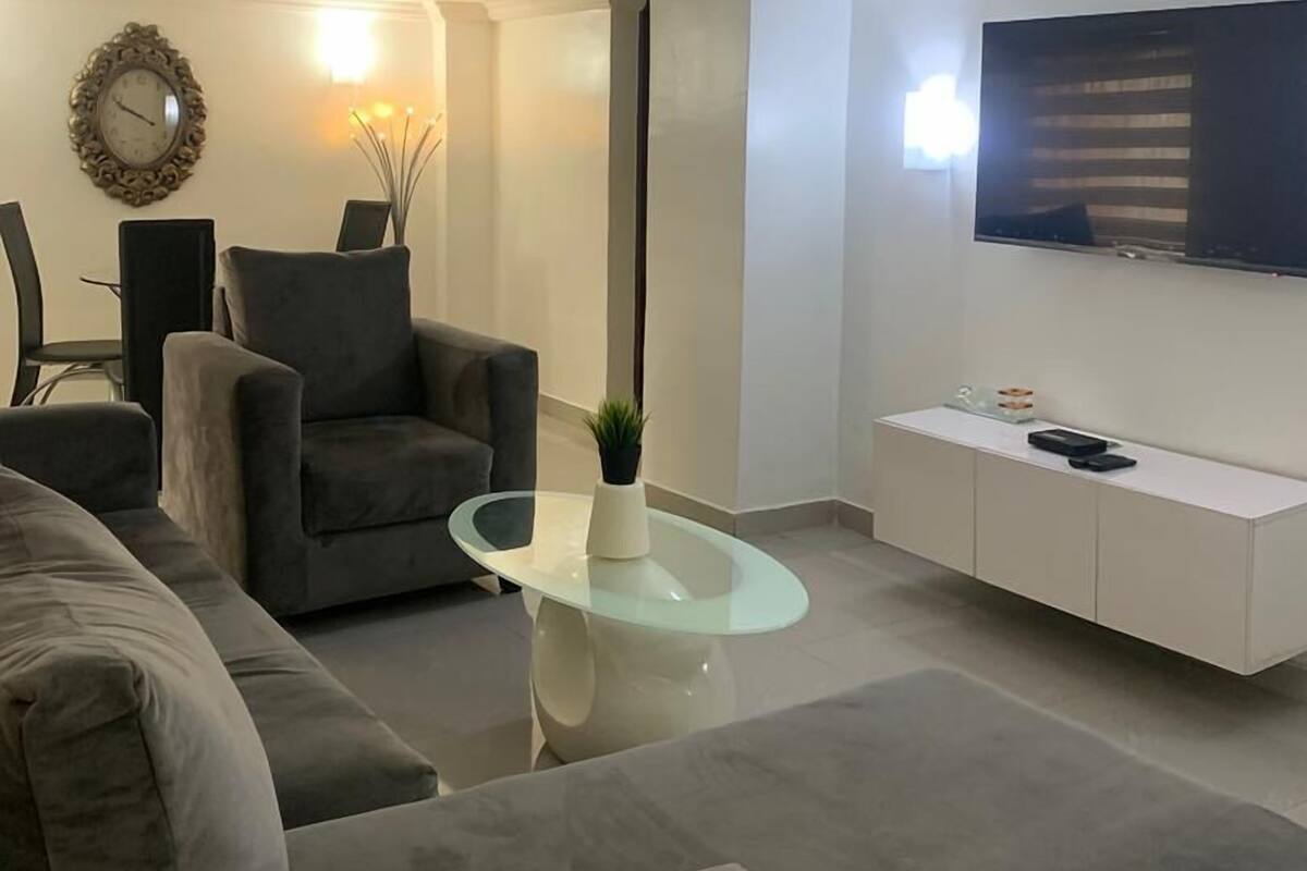 A modern living room with a grey sofa, a glass coffee table, a grey armchair, a white cabinet, and a flat-screen TV mounted on the wall.