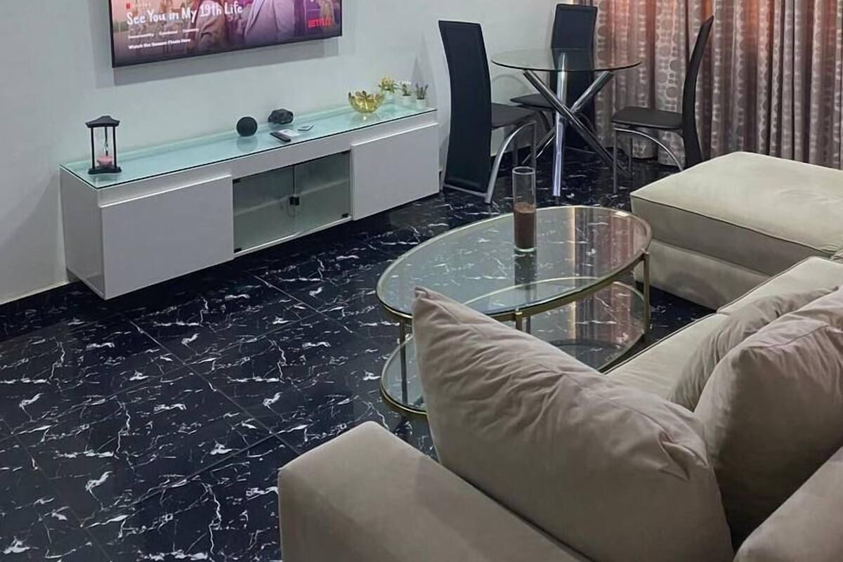 A modern living room with a flat-screen TV, a glass coffee table, and beige sofas.