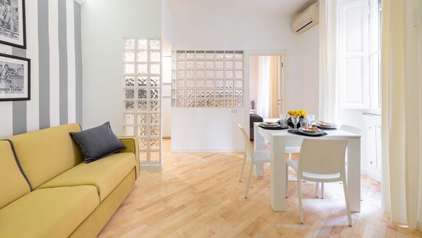 A modern living room with a yellow sofa, a dining table with white chairs, and a decorative glass partition. - Cozy apartment 5 minutes walk from the Colosseum (Roma)