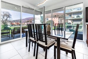 Een eetgedeelte met een glazen tafel en stoelen, grote ramen en uitzicht op gebouwen en heuvels.
