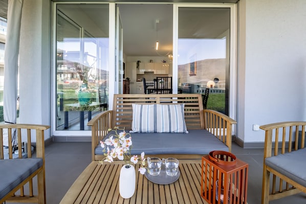 A balcony with a sofa, chairs, a table with a vase and glasses, and a view of the interior.