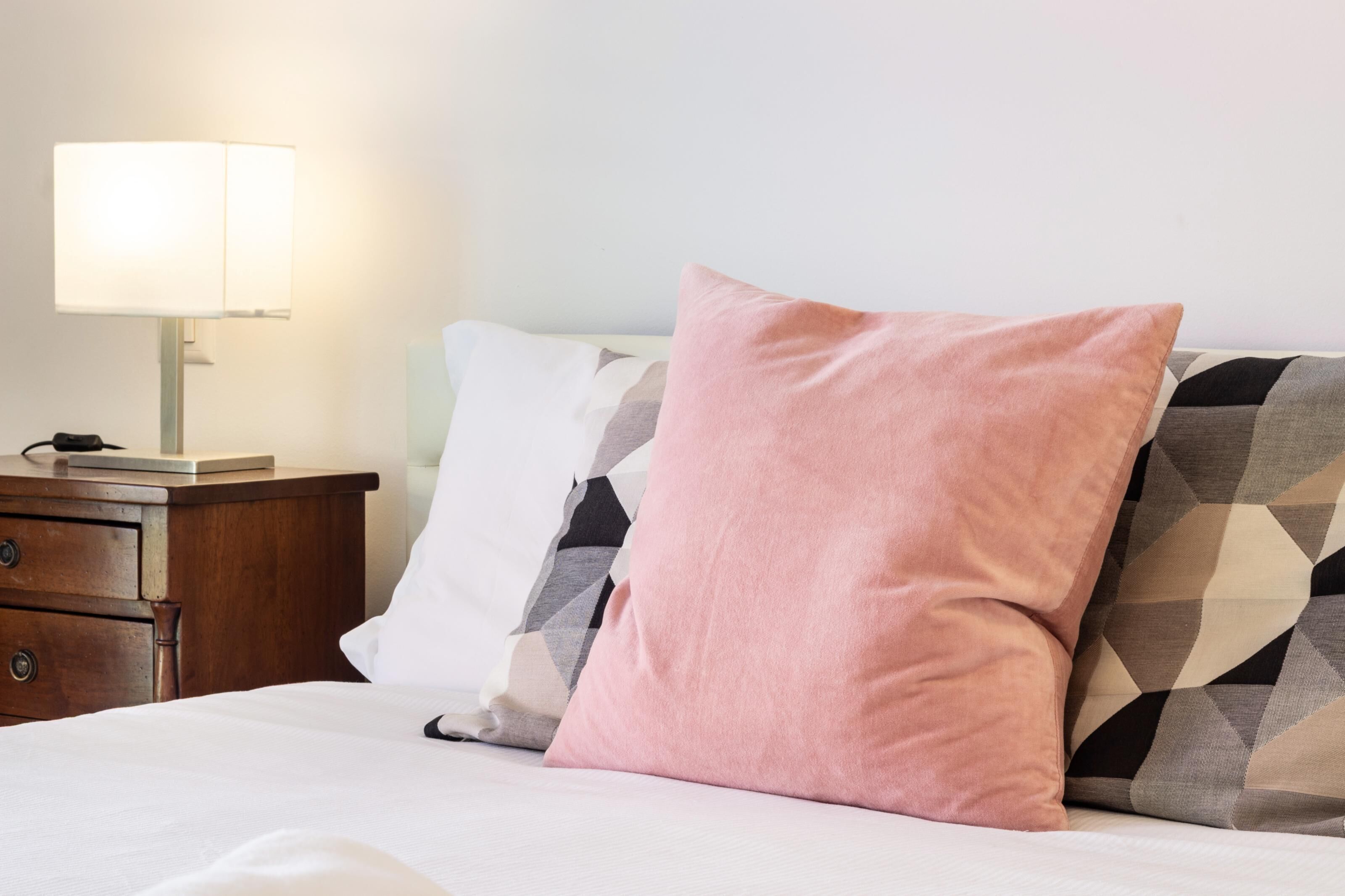 A bed with a white and patterned pillow, a bedside table with a lamp, and a wooden nightstand.