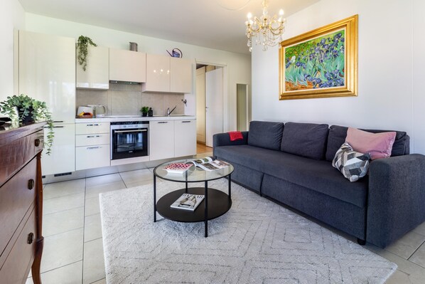 Een moderne woonkamer met een grijze bank, een ronde salontafel en een keuken op de achtergrond.