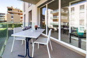 Een balkon met een tafel en stoelen, een glazen hekwerk en uitzicht op omliggende gebouwen.