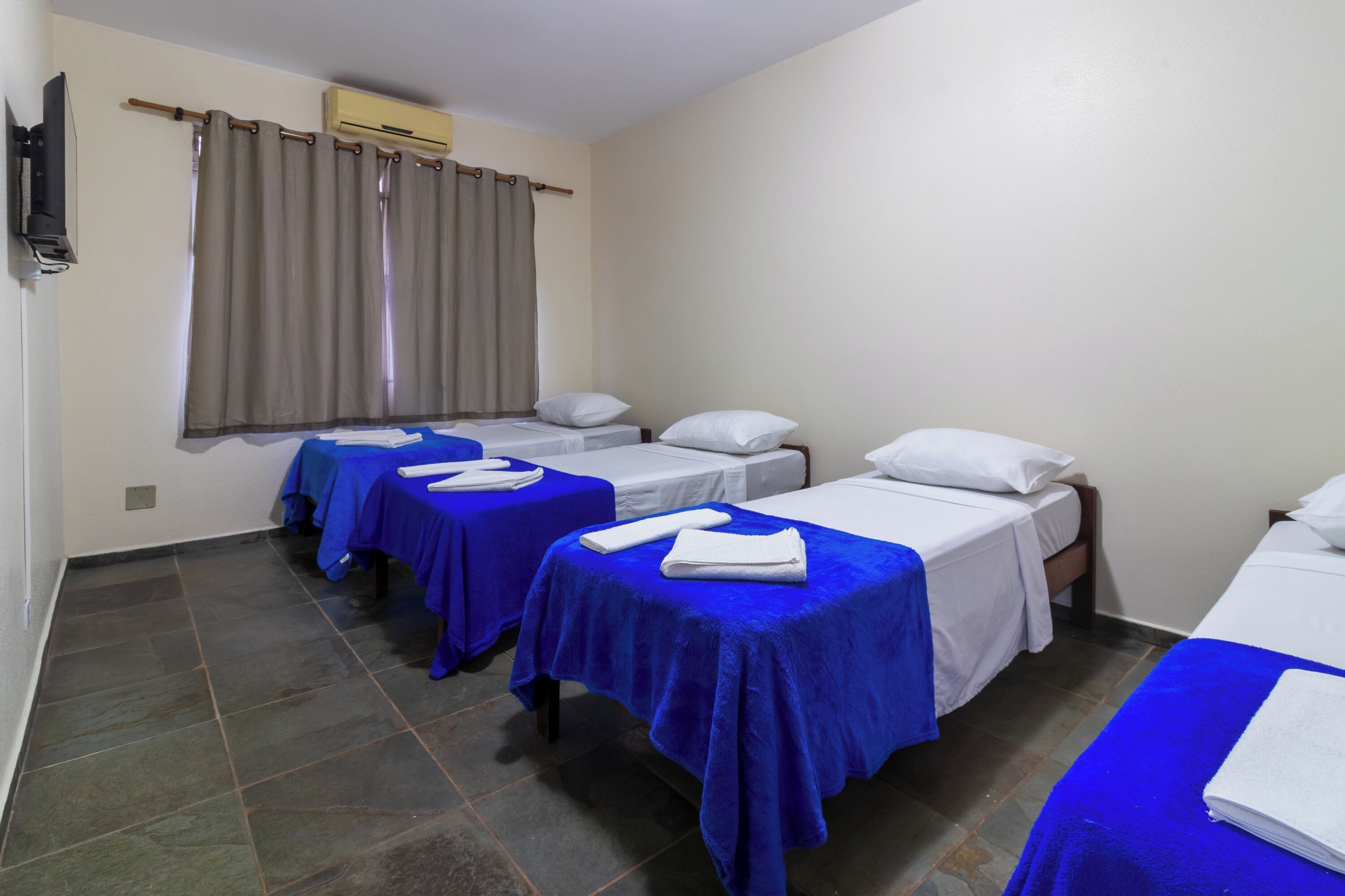 A hotel room with three single beds, each with a blue bedspread and white pillows. There is a window with curtains, a flat-screen TV mounted on the wall, and a small air conditioning unit above the window.