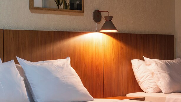 A neatly made bed with white bedding and a tan throw blanket, a wooden headboard, and a wall-mounted lamp. - Foz Central Budget Hotel (Foz do Iguacu)