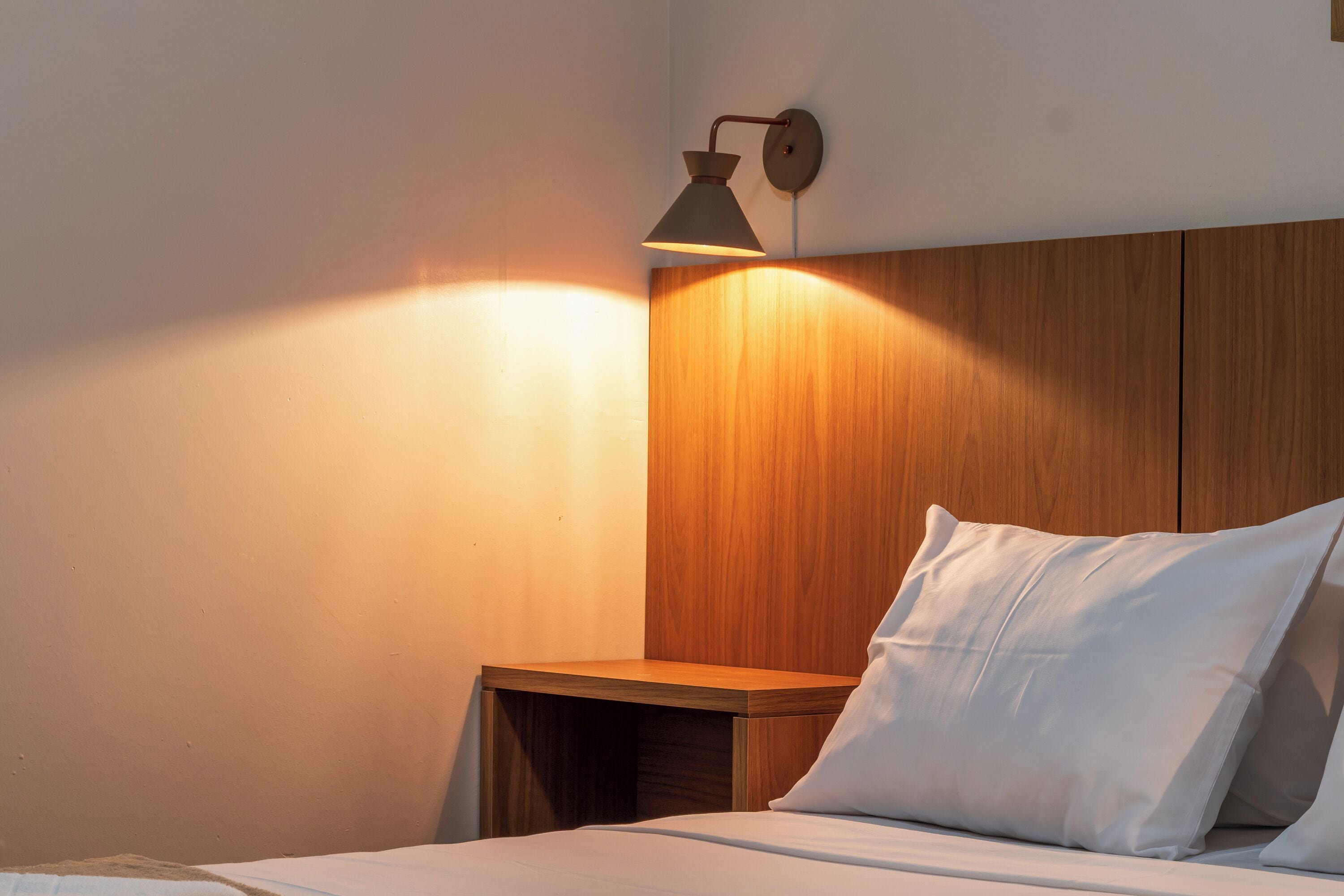 A neatly made bed with white linens and pillows, a wooden nightstand, and a wall-mounted lamp.