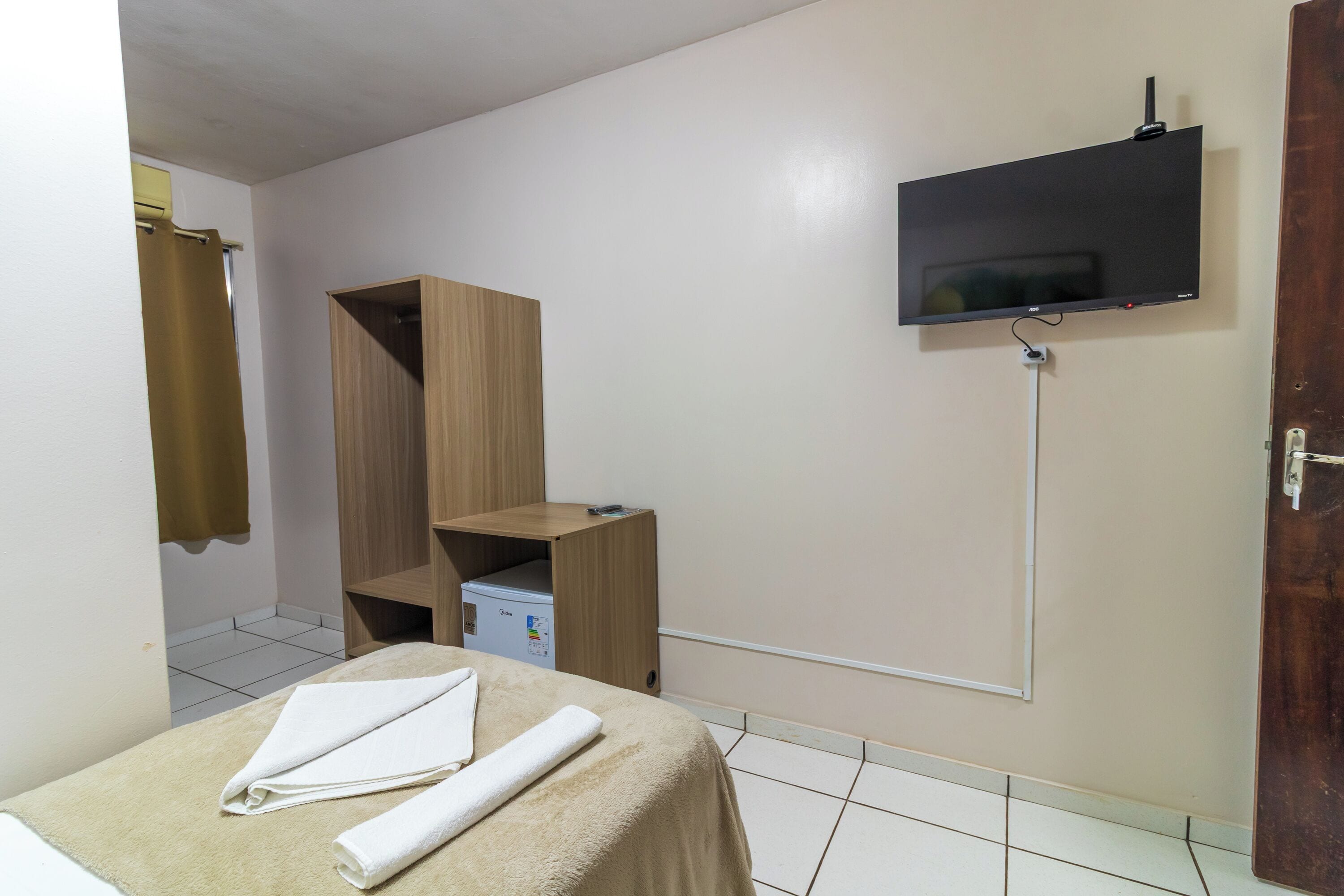 A hotel room with a bed, a TV mounted on the wall, a wooden wardrobe, and a small storage unit.