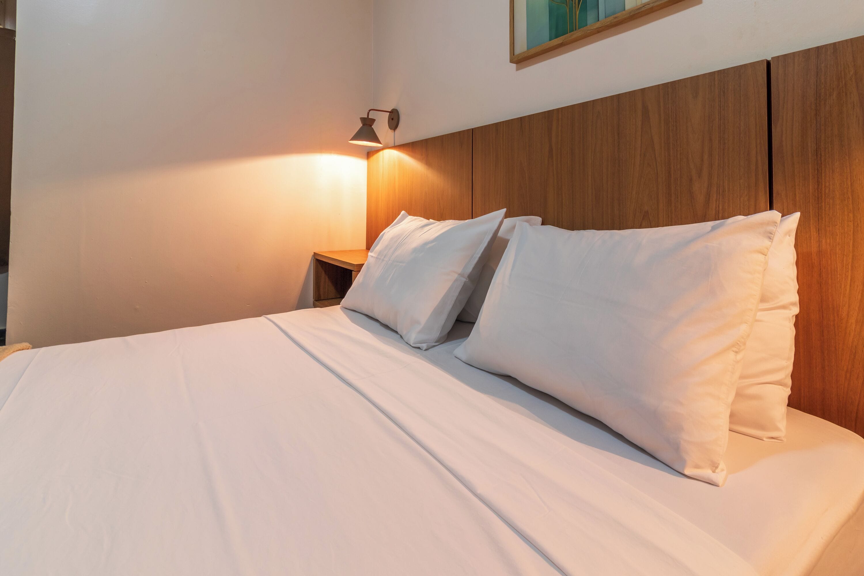 Ein ordentlich bezogenes Bett mit weißen Leinen und Kissen, ein Holz-Kopfstück sowie ein an der Wand befestigter Nachttisch mit Lampe.