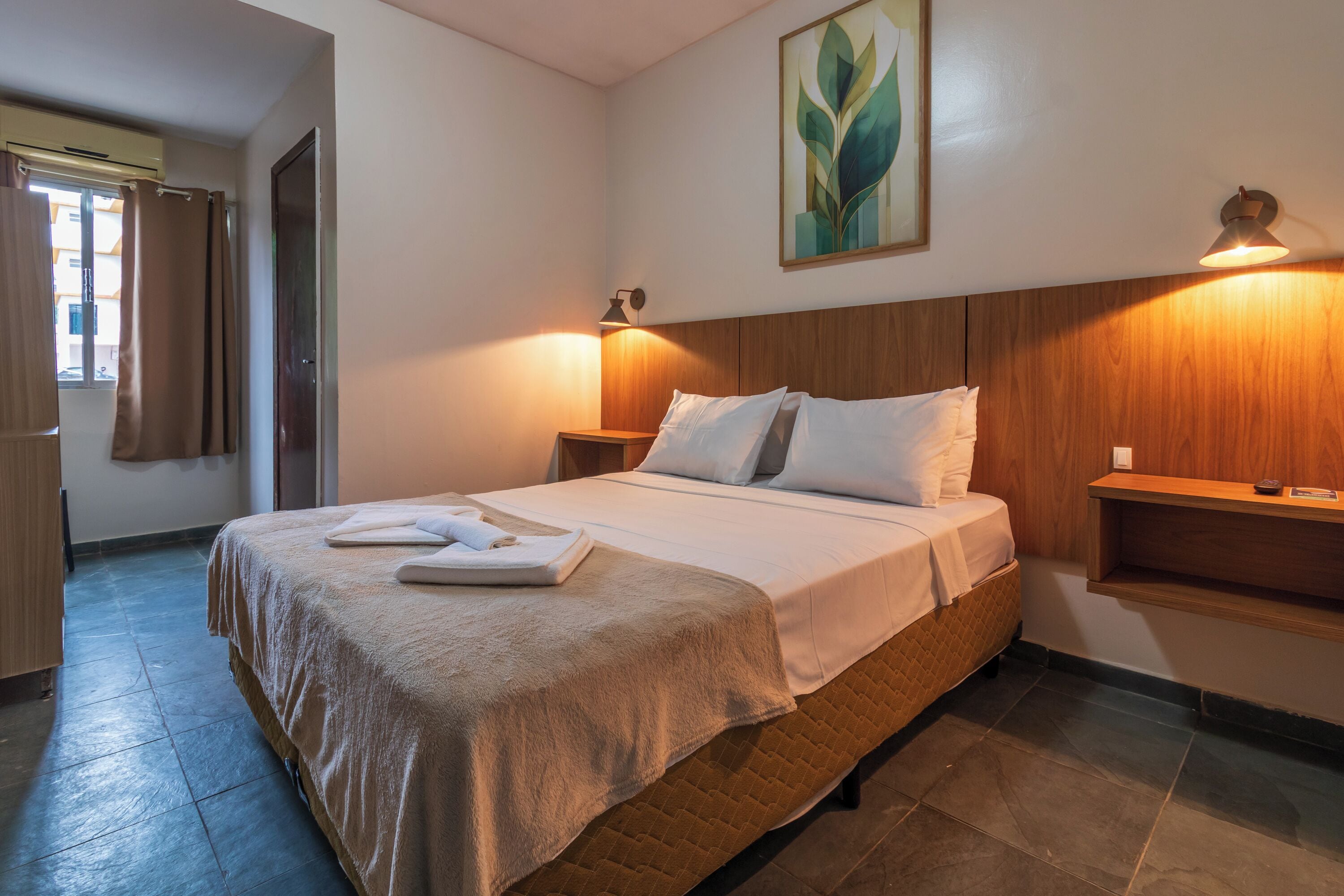 A neatly made bed with a quilted cover, a bedside table with a lamp, and a wall-mounted shelf.