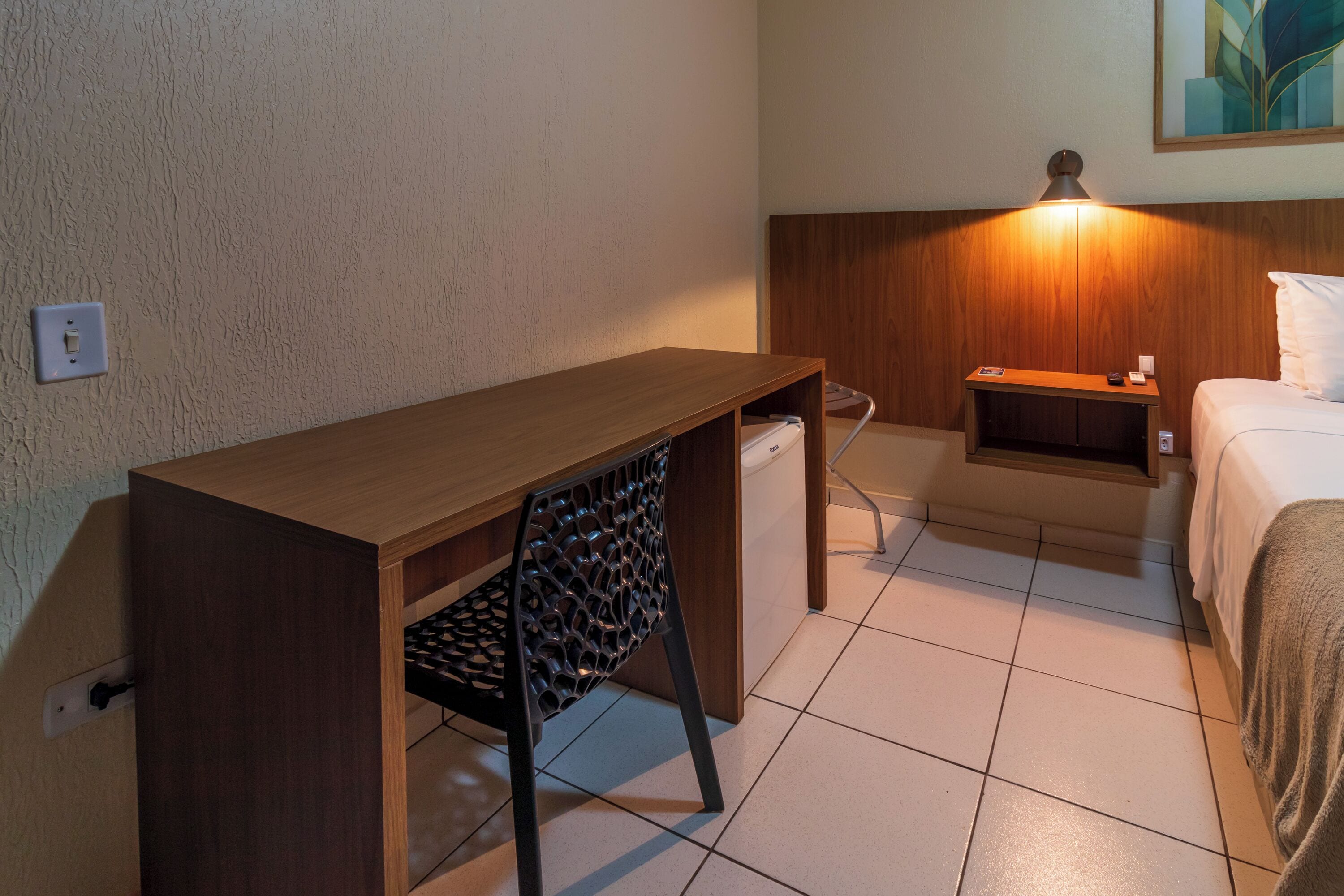 Ein Hotelzimmer mit einem Holztisch, einem Stuhl, einem Nachttisch mit Lampe, einem Bett und einem Wandbild.