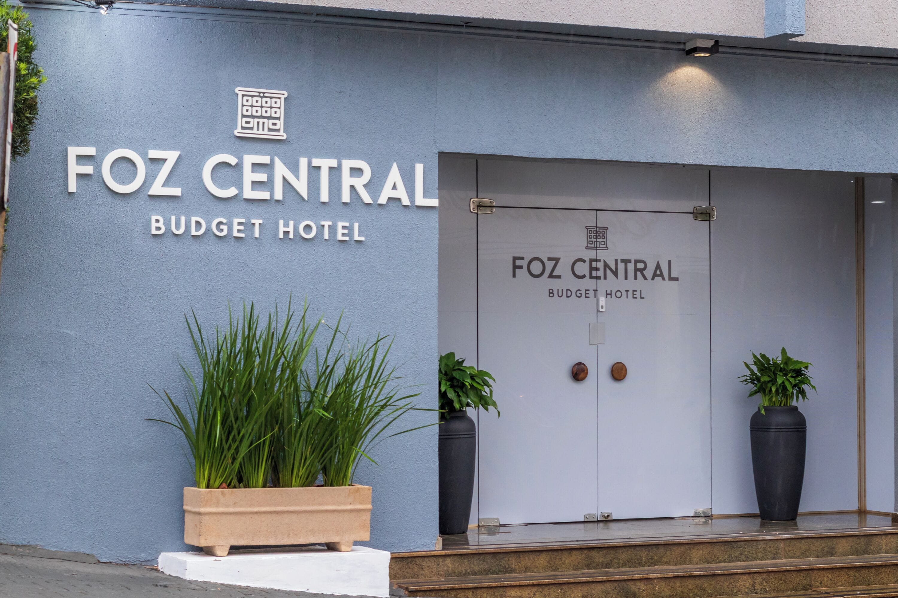 Foz Central Budget -hotellin ulkopuoli: moderni julkisivu, suuri sisäänkäyntiovi ja kaksi ruukussa olevaa kasvia molemmin puolin ovea.