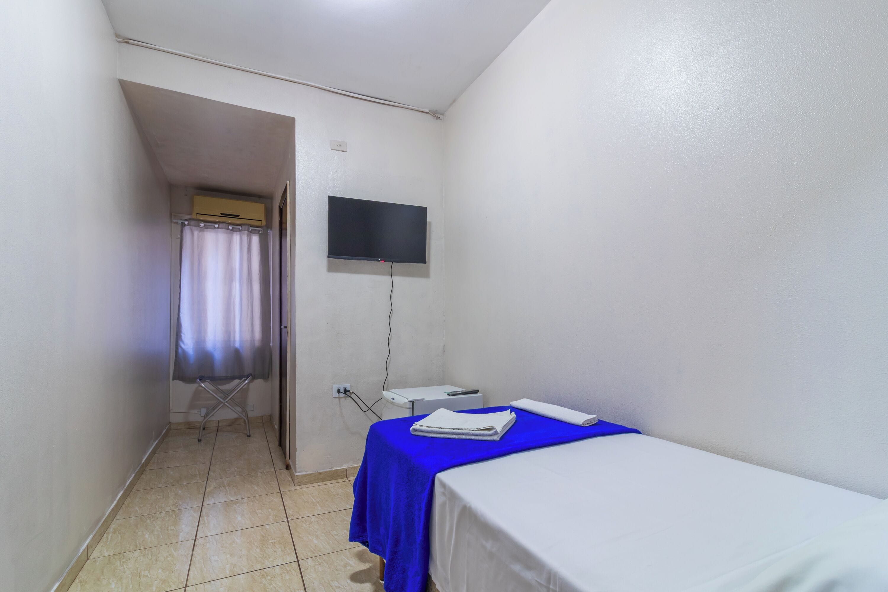 A small, clean room with a single bed, a flat-screen TV mounted on the wall, and a small table with a towel.