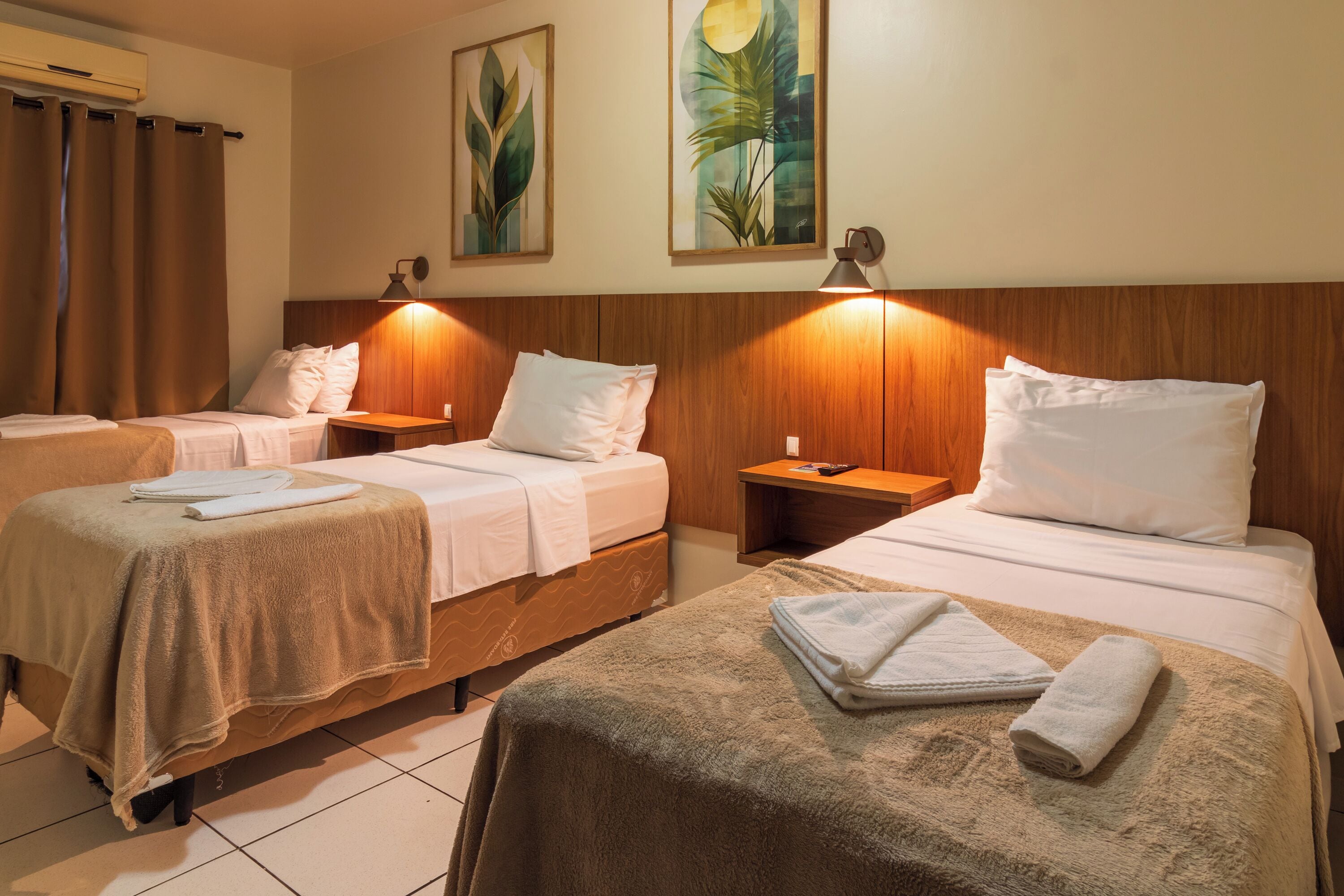 Habitación de hotel con dos camas individuales, cada una con cabecero de madera, mesita de noche y lámpara. Se observan dos cuadros colgados en la pared, una ventana con cortinas y suelo de baldosas.