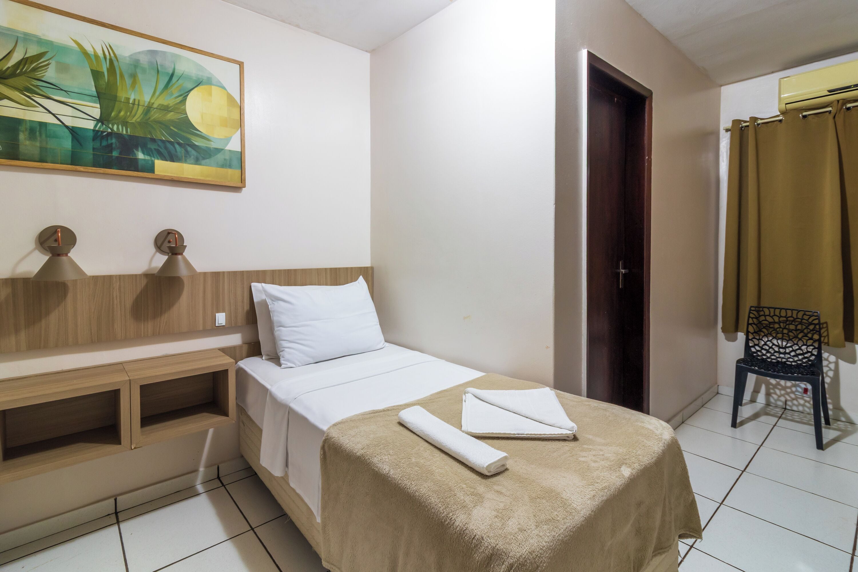 A hotel room with a bed, a wooden headboard, a painting, a desk with two lamps, a chair, and a door.
