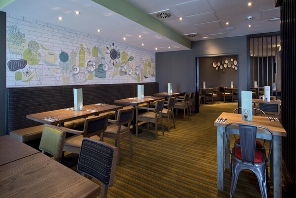 A restaurant with wooden tables and chairs, a brick wall mural, and a patterned carpet.