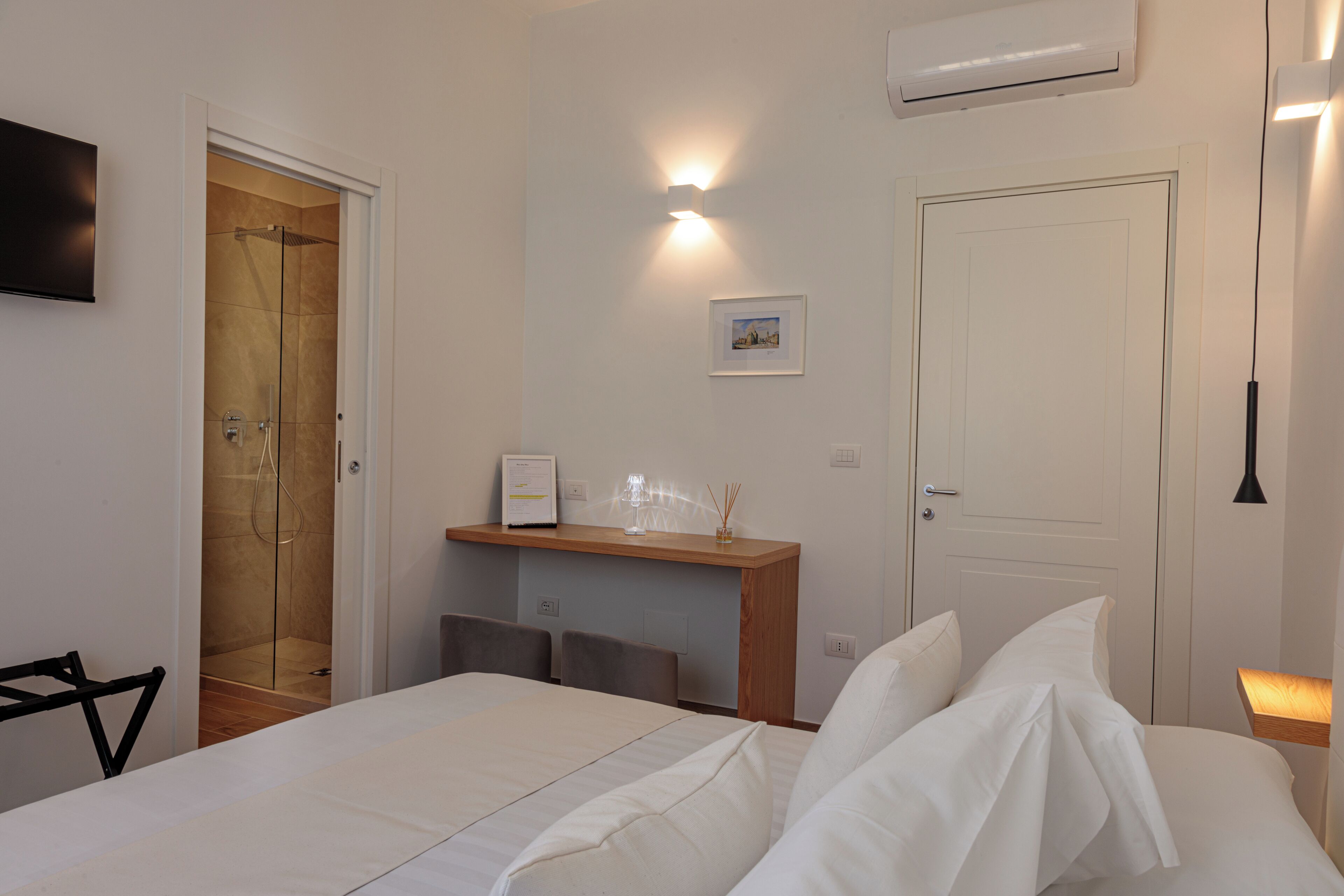A hotel room with a bed, a wooden nightstand, a framed certificate, a small shelf, a door, and a wall-mounted air conditioner.