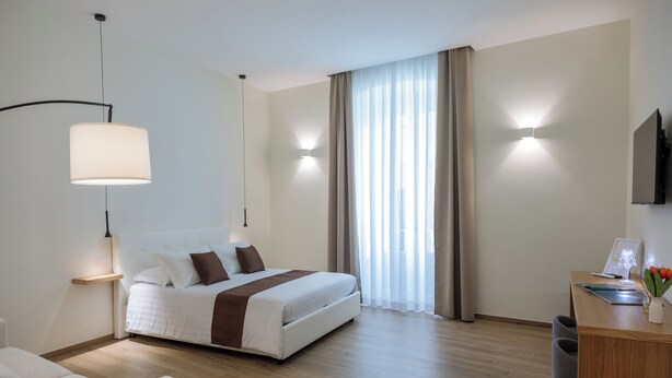 A modern hotel room with a bed, bedside table, lamp, and a desk with a chair. - Blanco Group (Trani)
