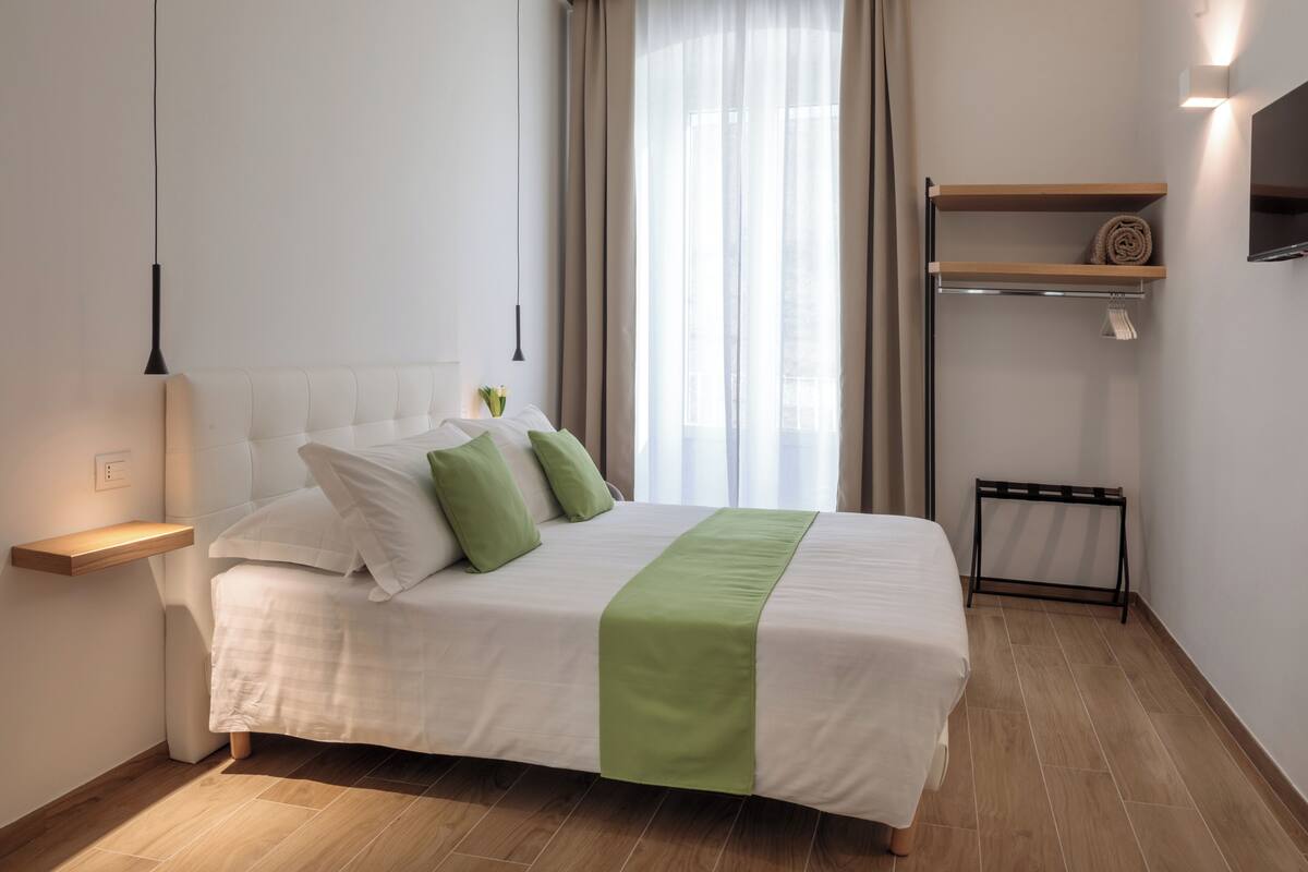 A neatly arranged bedroom with a bed, bedside tables, a TV, and a window with curtains.