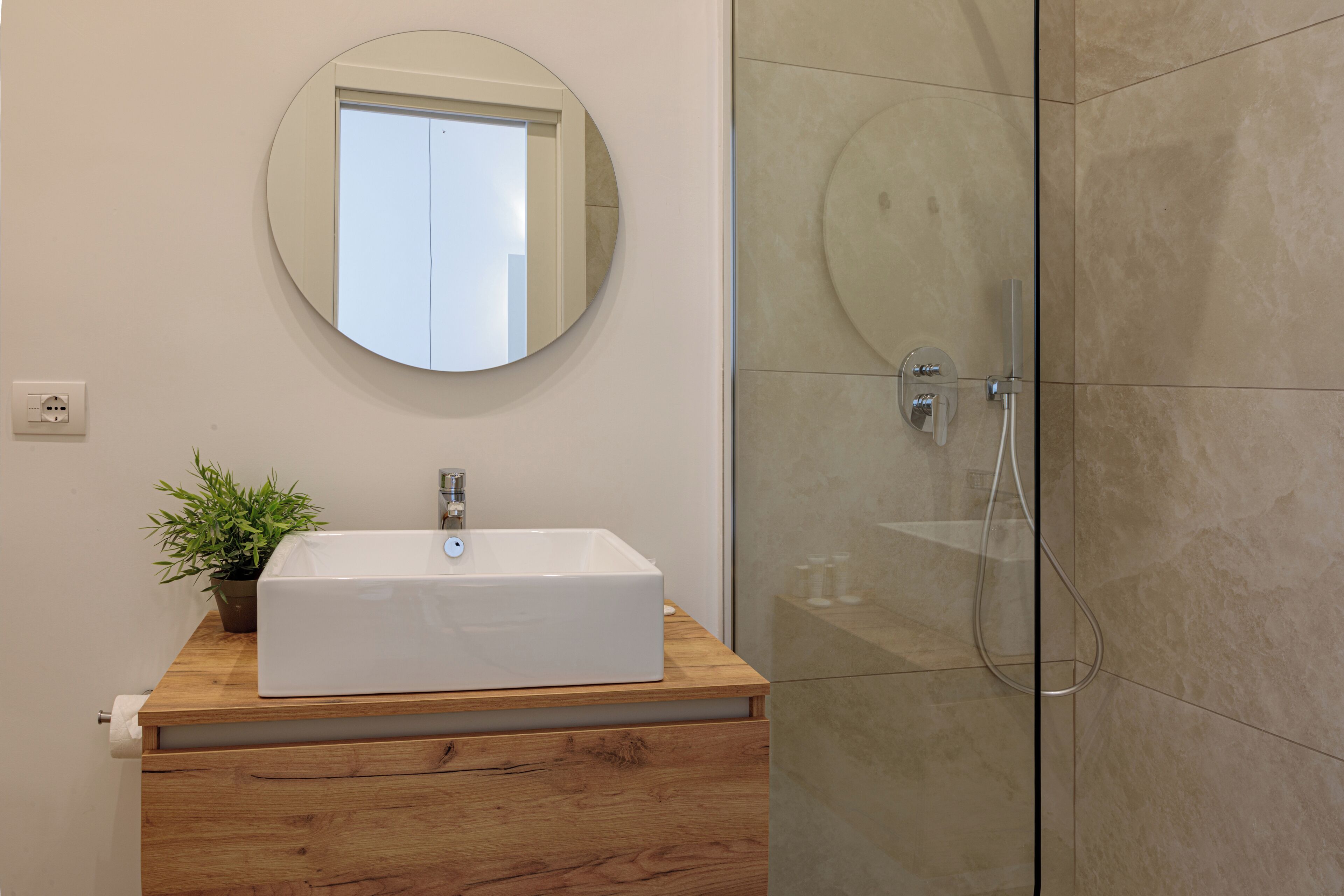 A bathroom with a white sink, wooden vanity, a round mirror, and a glass shower enclosure.