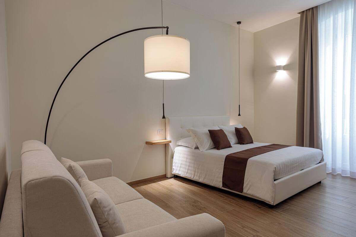 A modern hotel room with a bed, a sofa, a floor lamp, and a bedside table.