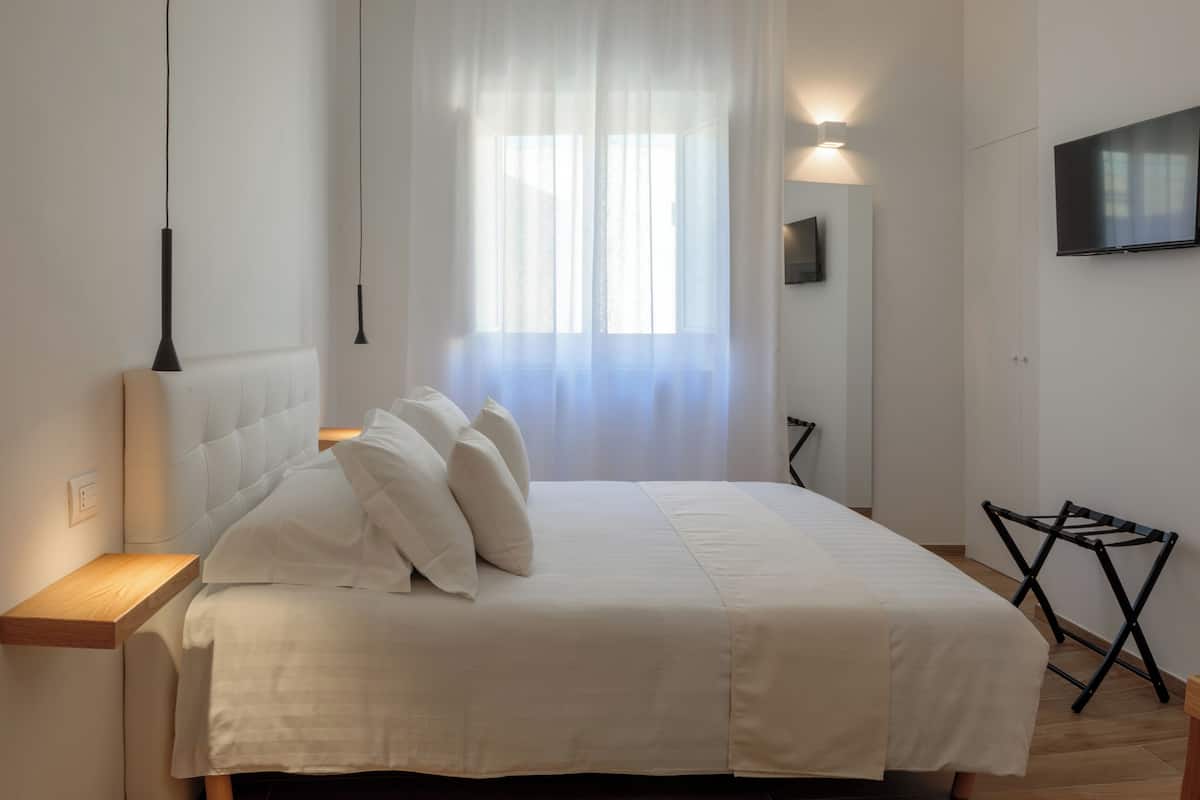 A neatly made bed with white linens and pillows, a bedside table with a lamp, a folding chair, and a TV mounted on the wall.