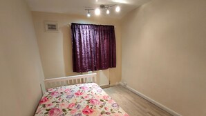 A small, single-room apartment with a bed, a radiator, a window with curtains, and a ceiling light fixture. - South London 3 Bedroom House (Carshalton)