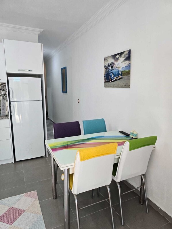 A compact kitchen with a refrigerator, a dining table with colorful chairs, and a painting on the wall.
