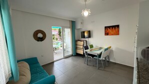 A living room with a sofa, a dining table with chairs, a TV, and a sliding glass door leading to an outdoor area. - Elfida Apart Palamutbükü (Datça)