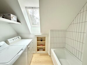Ein kleiner Hauswirtschaftsraum mit Waschmaschine, Wäscheständer und einer kleinen Ablagefläche. Ein Oberlicht lässt natürliches Licht herein, und ein gefliester Duschbereich ist sichtbar.