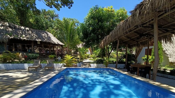 A rectangular swimming pool with a blue tiled interior, surrounded by a concrete deck. There are wooden chairs and tables under a thatched roof structure. - eco hotel amanecer tayrona (Santa Marta)