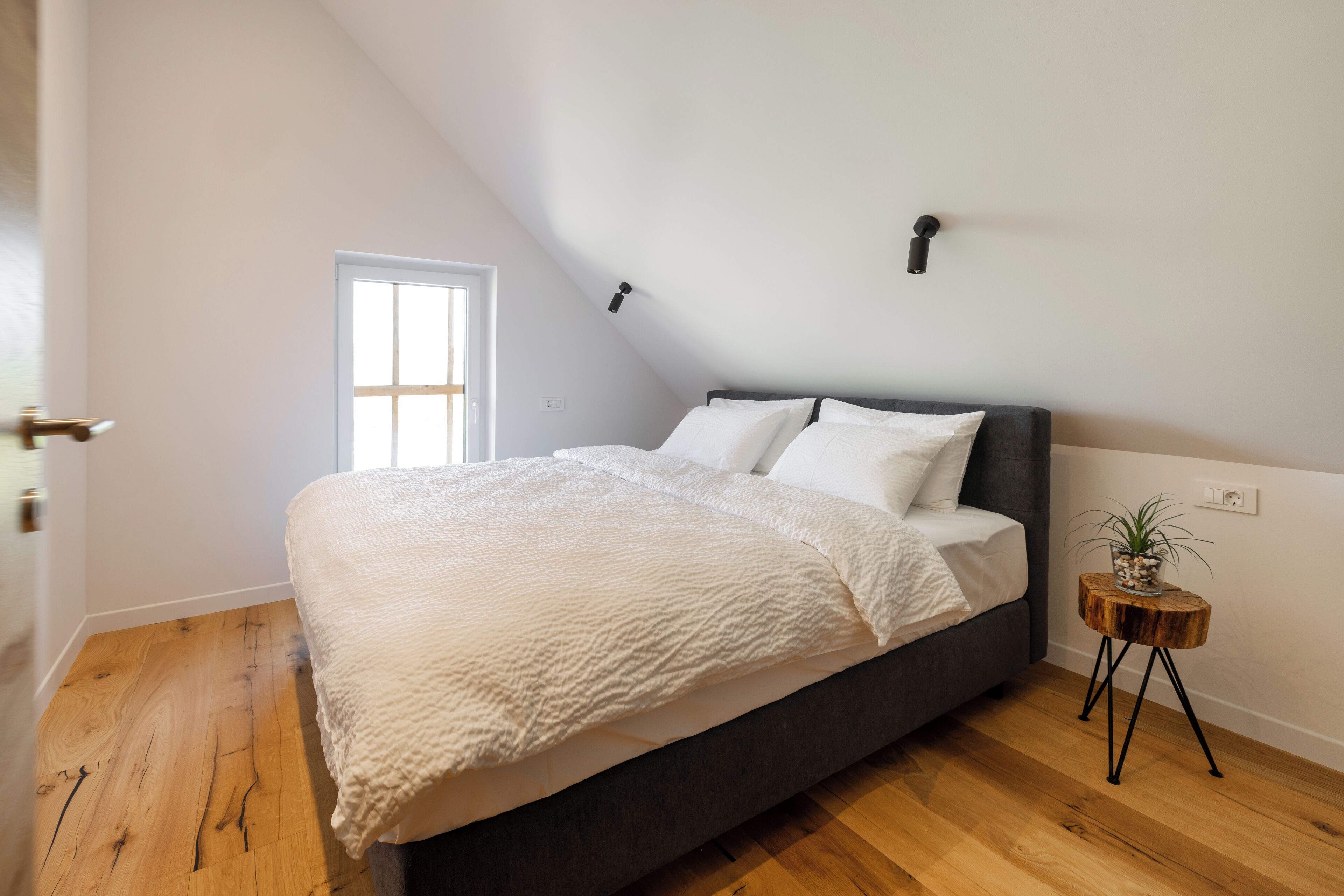 Ein Schlafzimmer mit einem Bett, einem kleinen Holfnitztisch mit einer Pflanze und einem Fenster.