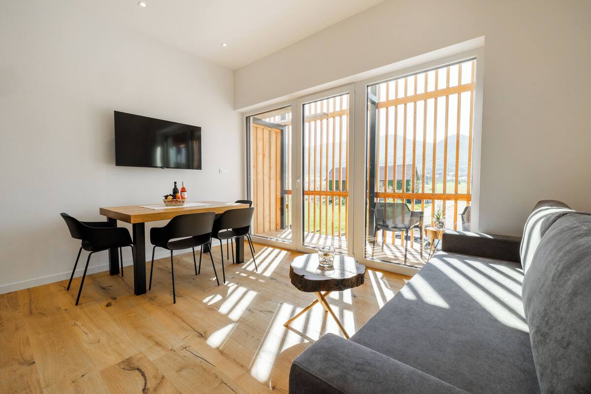 A modern living room with a wooden dining table, black chairs, a grey sofa, and a TV mounted on the wall.