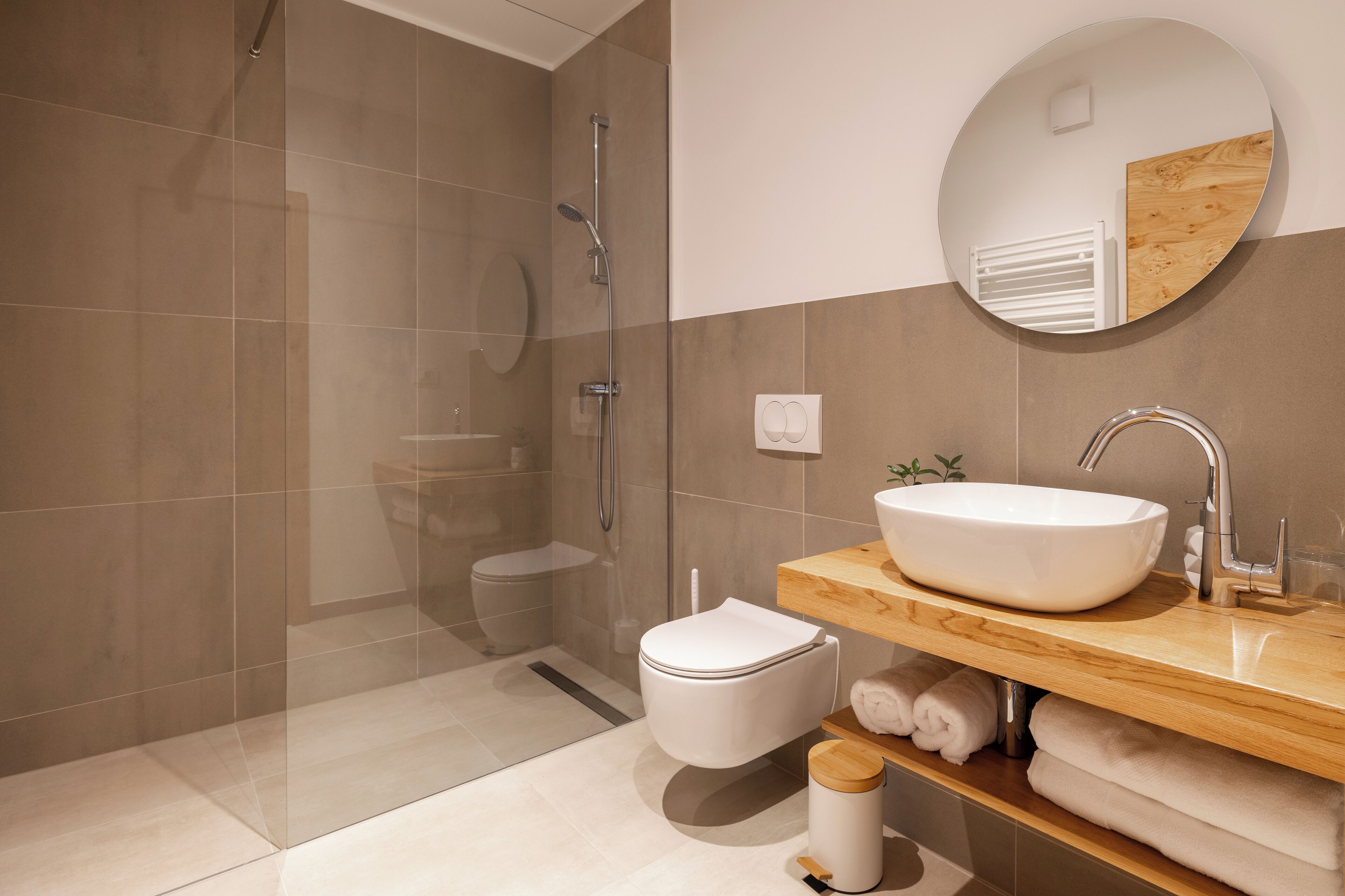 A modern bathroom with a glass-enclosed shower, a white toilet, a wooden vanity with a large sink, and a round mirror.