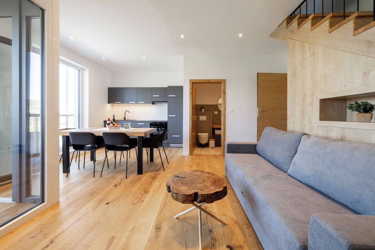 A modern kitchen with dark cabinets, a dining area with a wooden table and chairs, and a grey sofa in a living area.