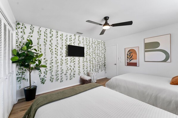 Un dormitorio con dos camas, un ventilador de techo, un televisor y una pared decorada con plantas colgantes.