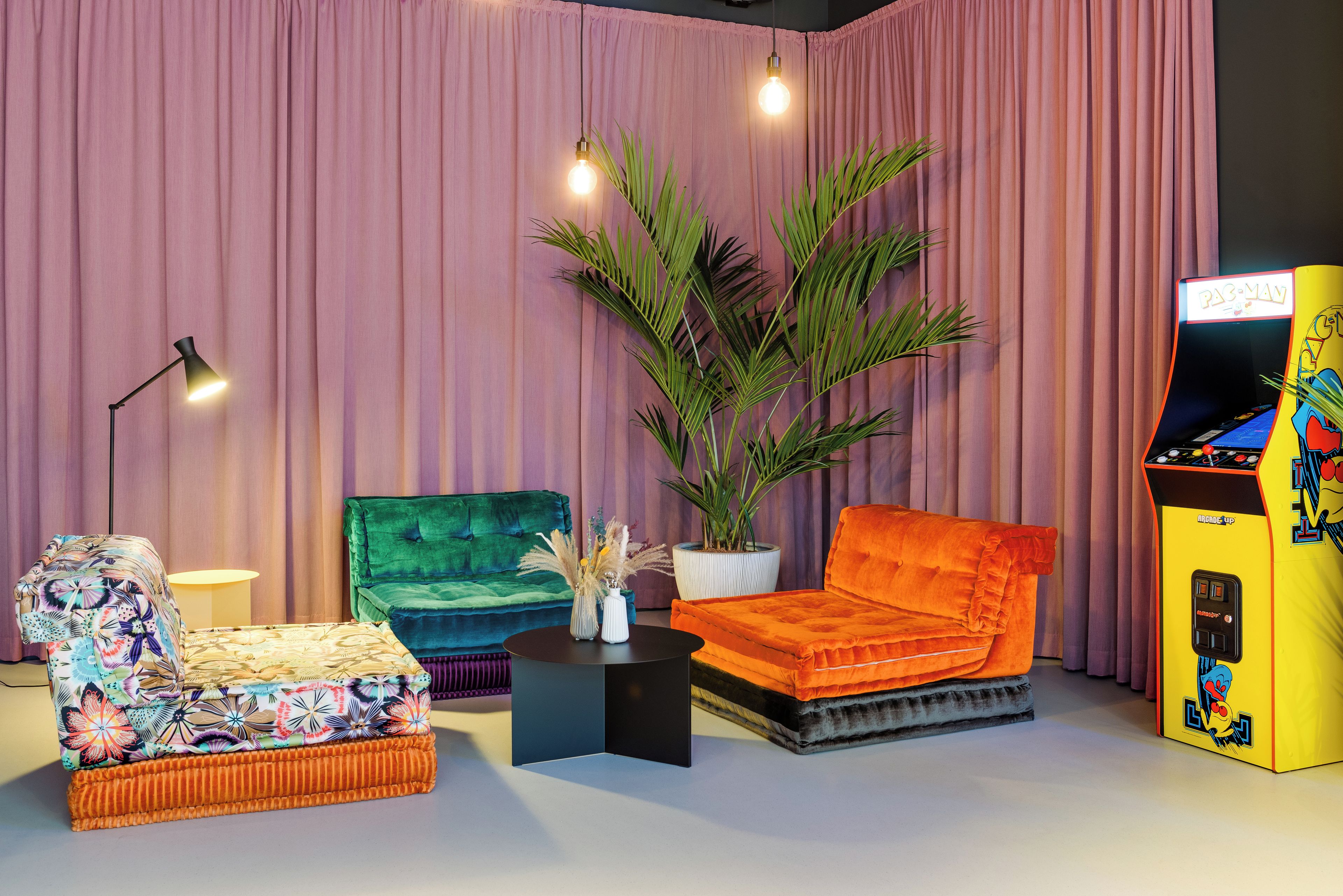 A room with a green sofa, an orange sofa, a patterned armchair, a potted plant, a lamp, and a Pac-Man arcade machine.