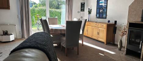 Ein gemütliches Wohnzimmer mit Kamin, einem Essbereich mit Tisch und Stühlen und Blick auf den Garten durch große Fenster.