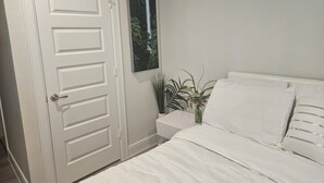 A bedroom with a white door, a bed with white bedding, a nightstand, a green plant, and a framed picture on the wall. - Luxury studio with AC, WiFi in wonderful Houston (Houston)