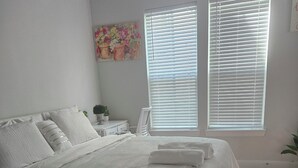 A bedroom with a bed, two windows with blinds, a nightstand, a potted plant, and a painting on the wall. - Luxury studio with AC, WiFi in wonderful Houston (Houston)