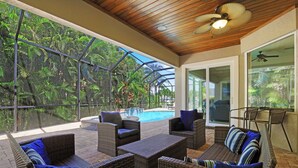 A covered outdoor area with wicker furniture, a pool, and a view of lush greenery.