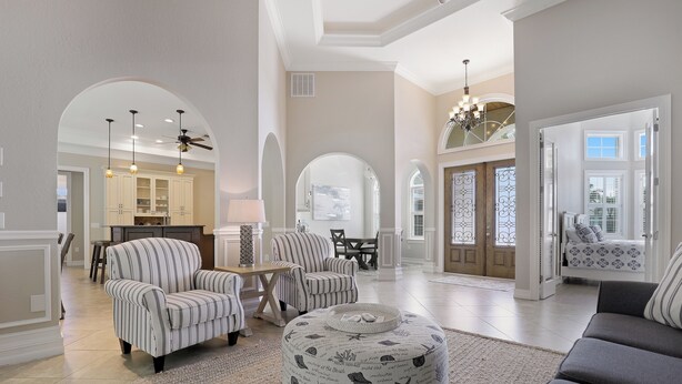 A spacious living room with a round ottoman, striped armchairs, and a grey sofa. There is a kitchen in the background with a dining area.