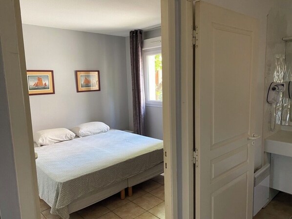 A bedroom with a bed, two framed pictures on the wall, a window with curtains, and a door with a telephone.